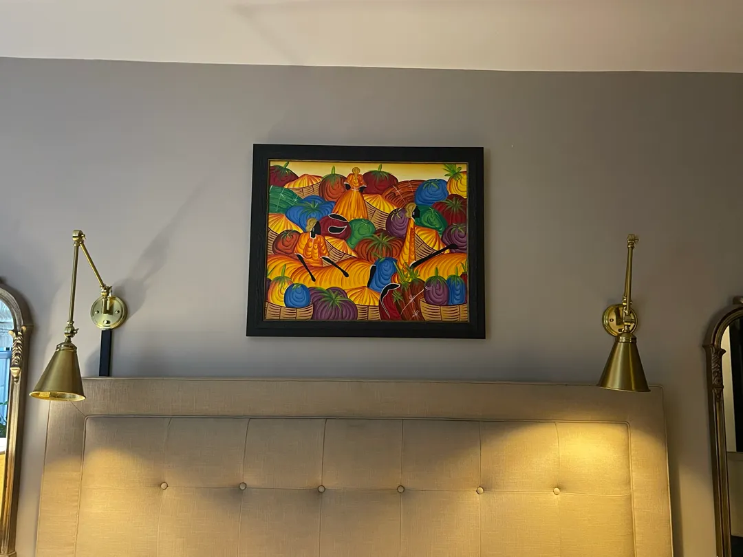 A black-framed colourful painting hanging above a tufted headboard between two brass swing-arm wall lights.