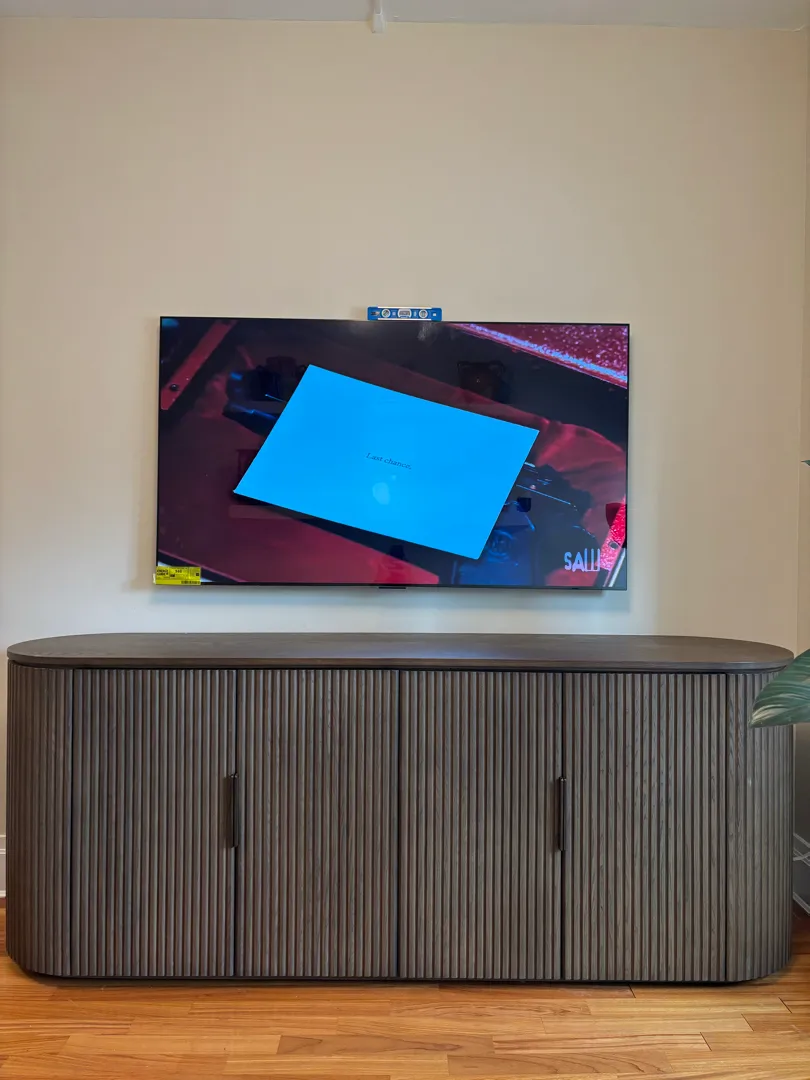 A TV mounted flat on a cream wall above a long dark ribbed console, with a small level resting on top.