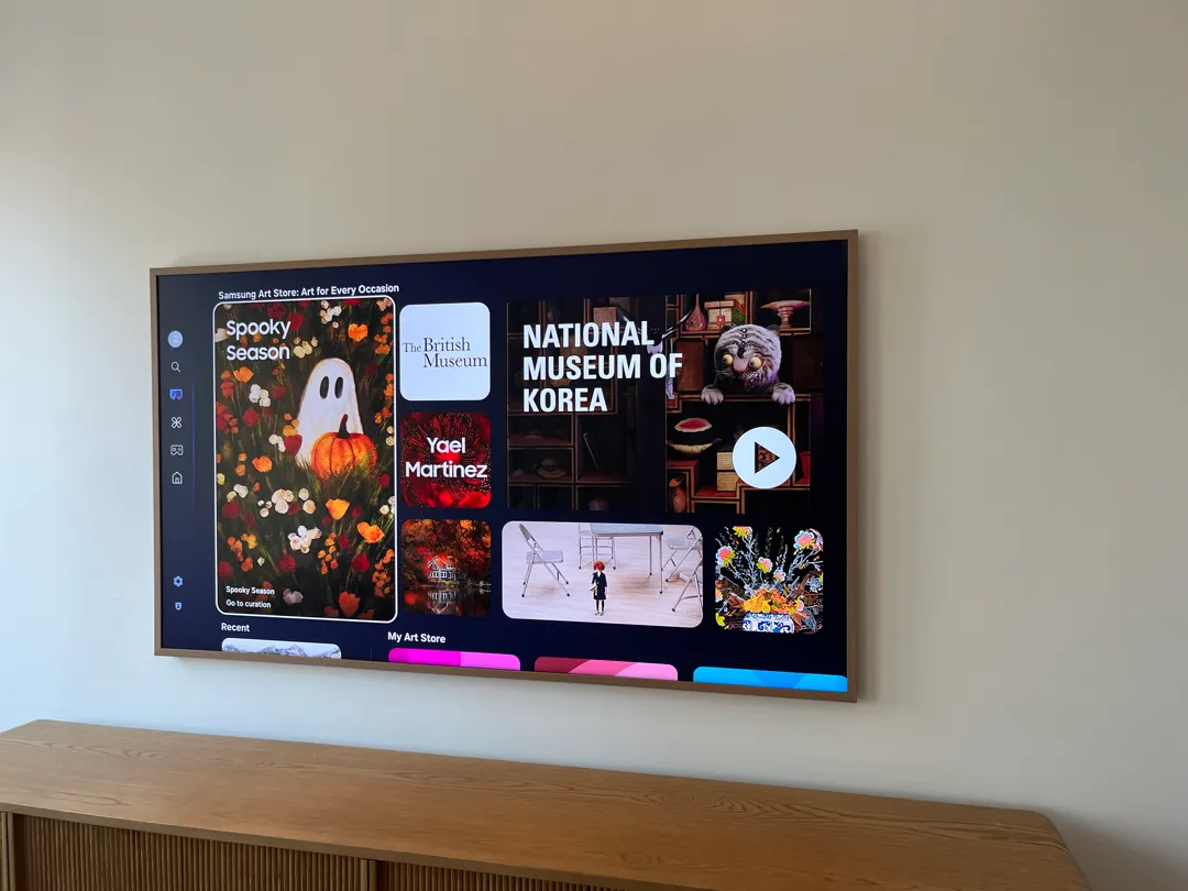 A wood-framed TV mounted on a wall shows an art store browsing menu, above a wooden console.