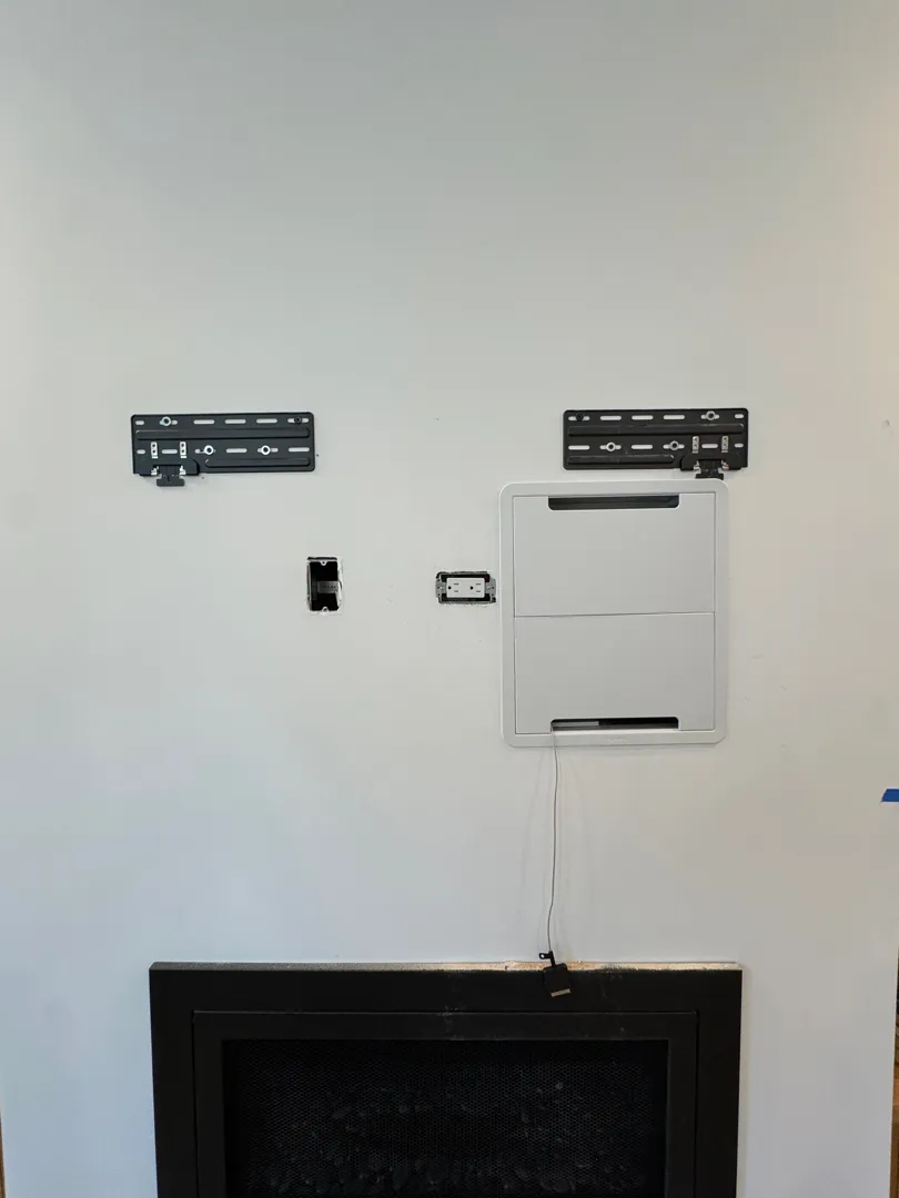 Closed recessed AV box and black mount plates installed above a black fireplace.