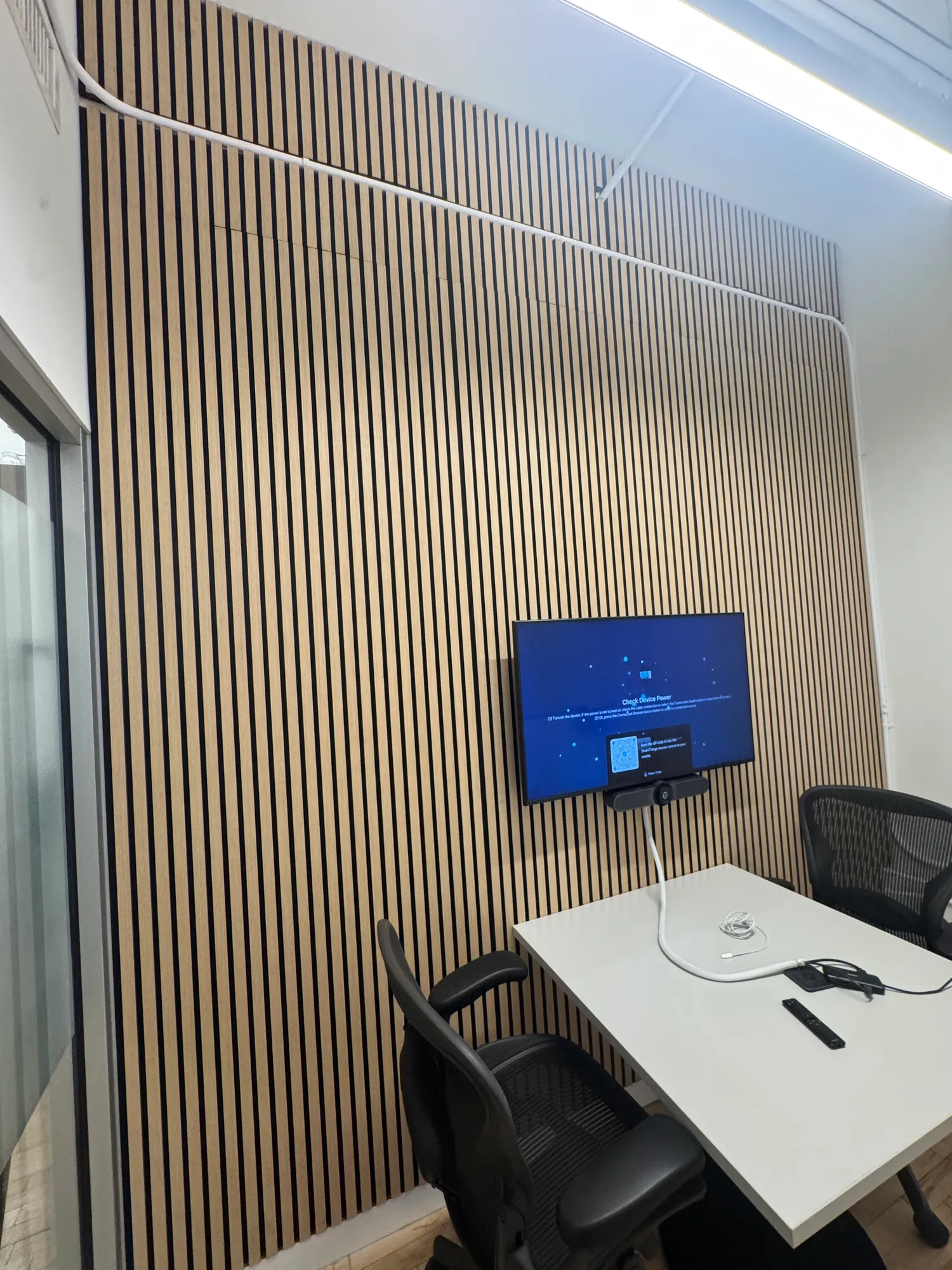 A small two-person meeting room with a TV mounted on a wood panel wall, showing a device power-check screen.