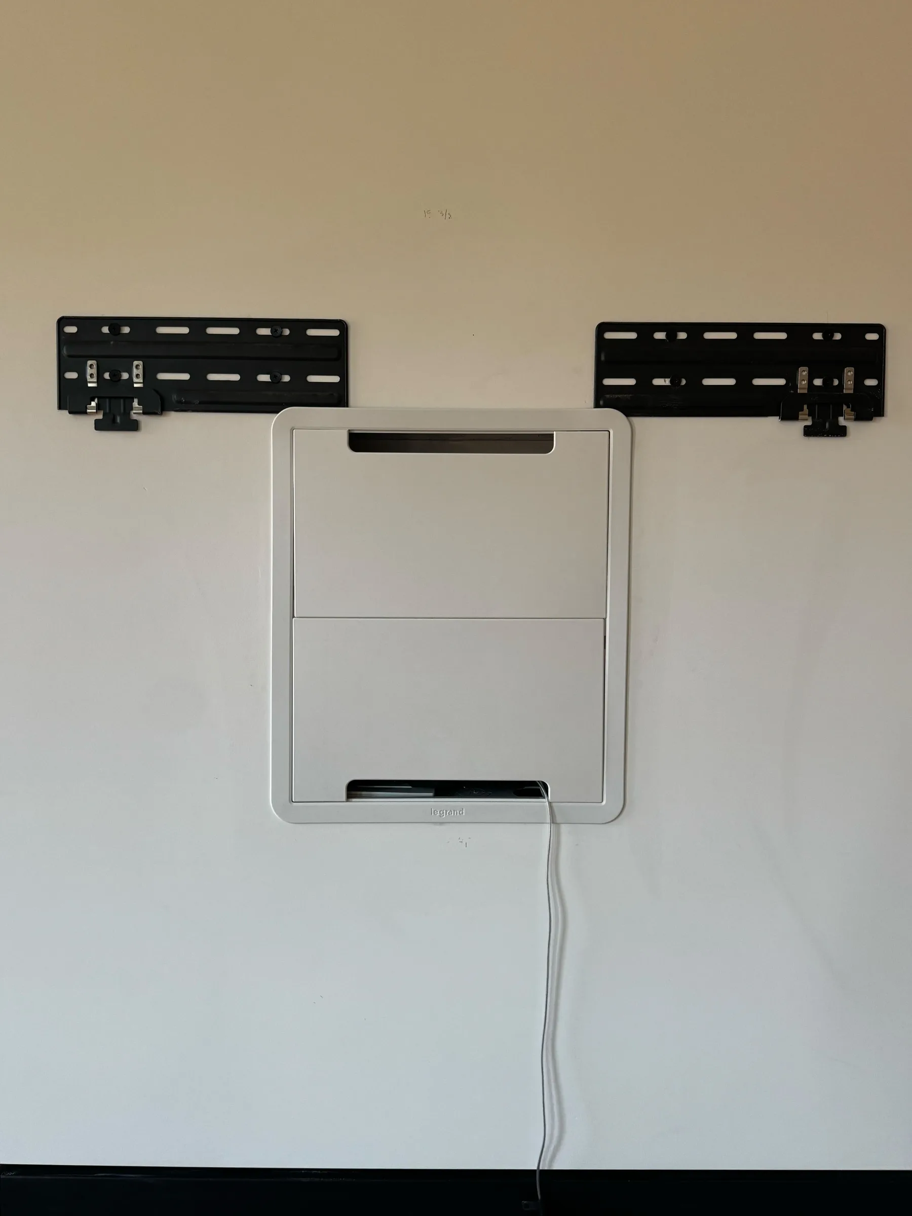 A closed white in-wall AV box between two black TV mount brackets on a wall, with a cable hanging below.