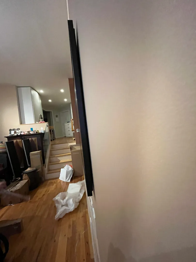 The TV's edge mounted flush to the wall, a cable running up toward the ceiling, with moving boxes nearby.