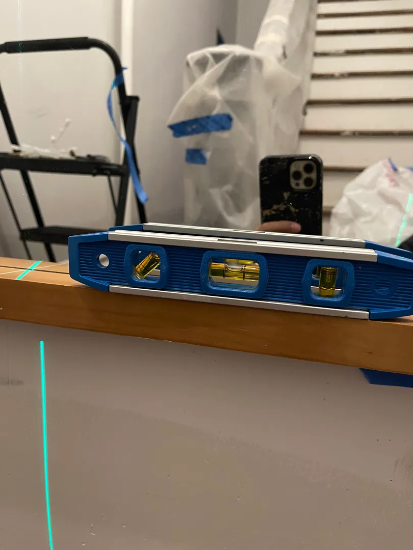 A level tool with laser guide lines, resting on a shelf near a plastic-wrapped staircase.