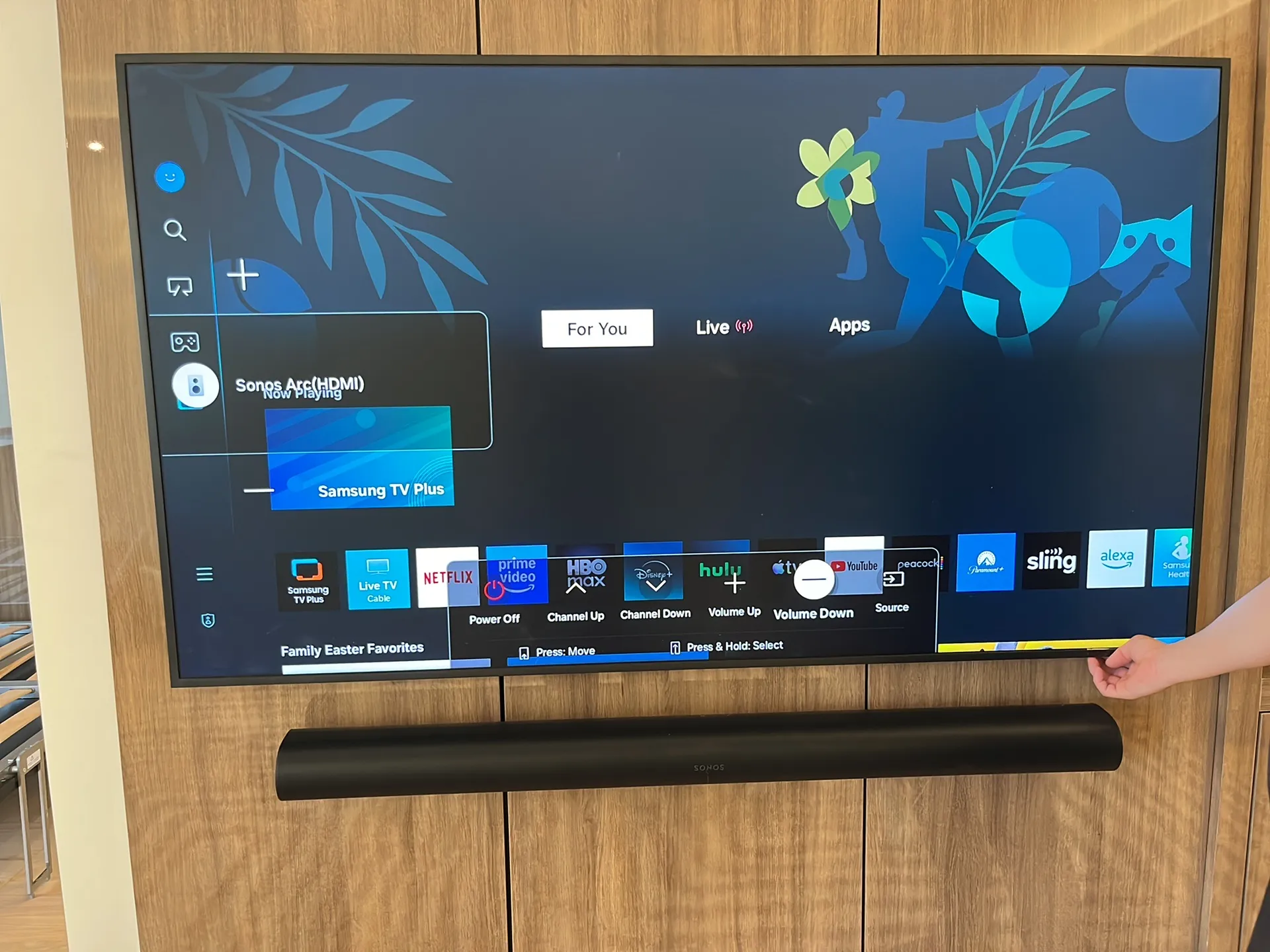 A hand reaches toward a wall-mounted TV showing an on-screen remote control overlay, with a soundbar mounted below.