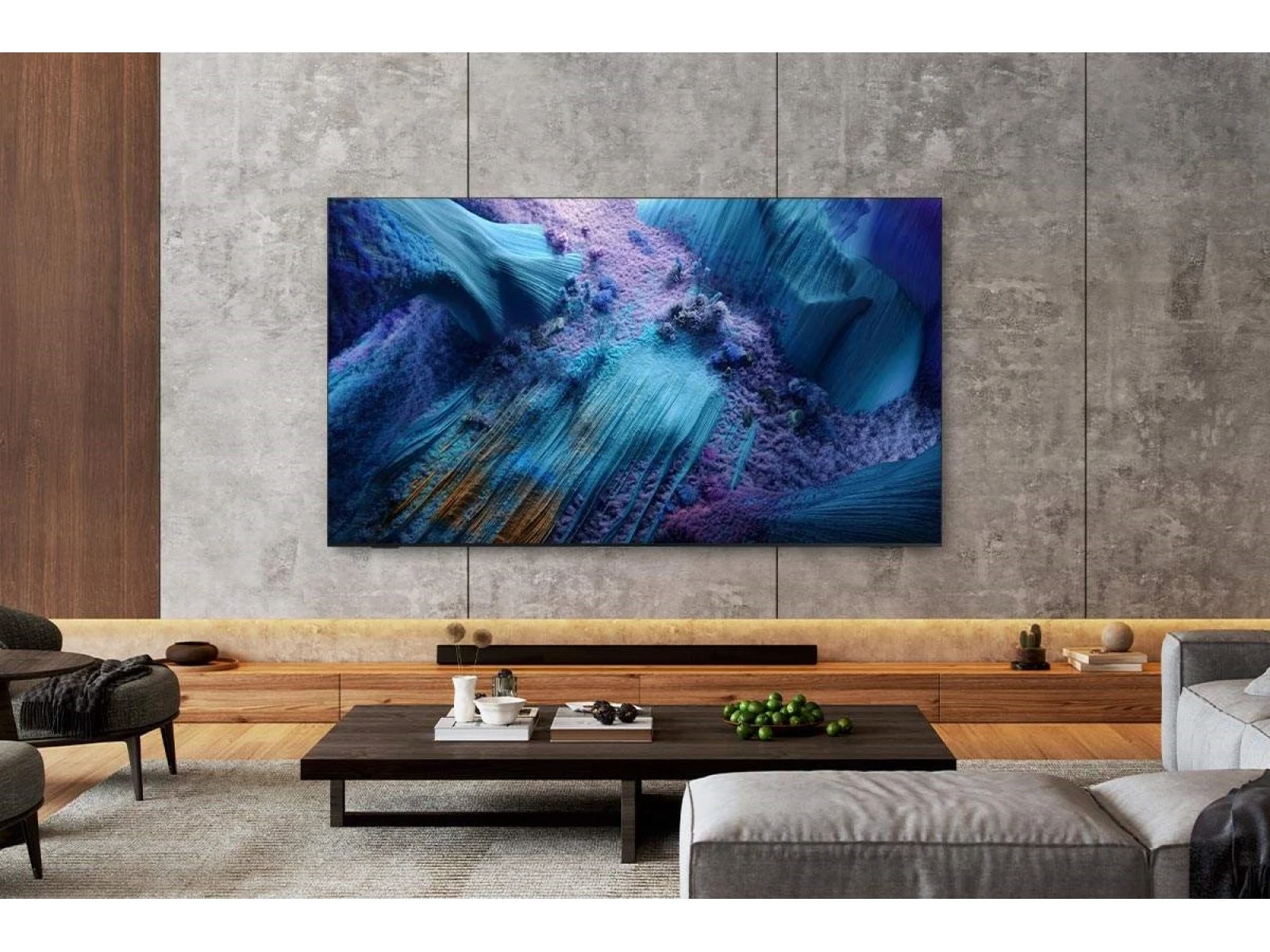 The QN990F wall-mounted in a modern concrete-walled living room with a wood console, soundbar and potted plants, showing a blue and purple abstract image.