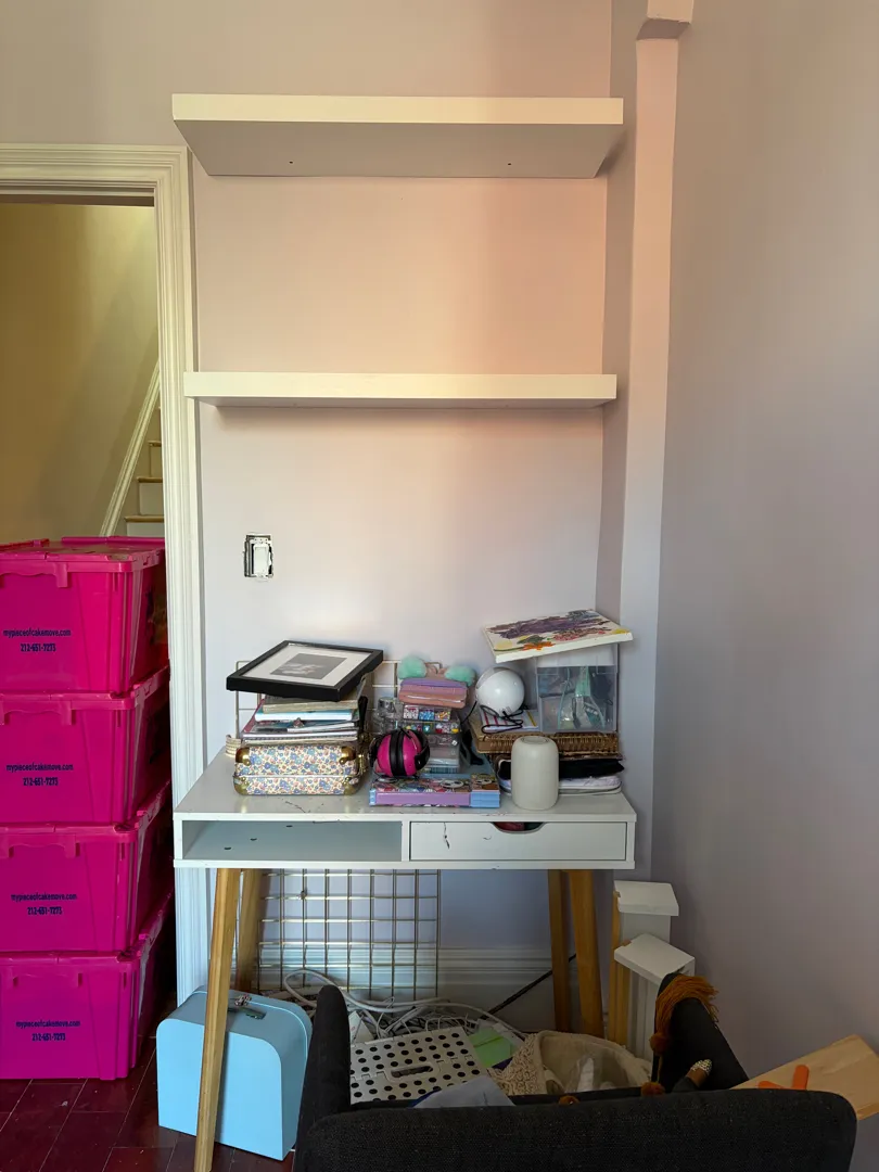 Two white floating shelves mounted above a small desk, moving bins visible nearby.