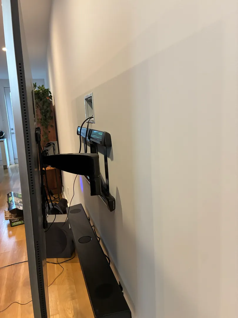 TV on a full-motion wall mount arm is extended out from the wall, with a round speaker and a soundbar on the floor and.
