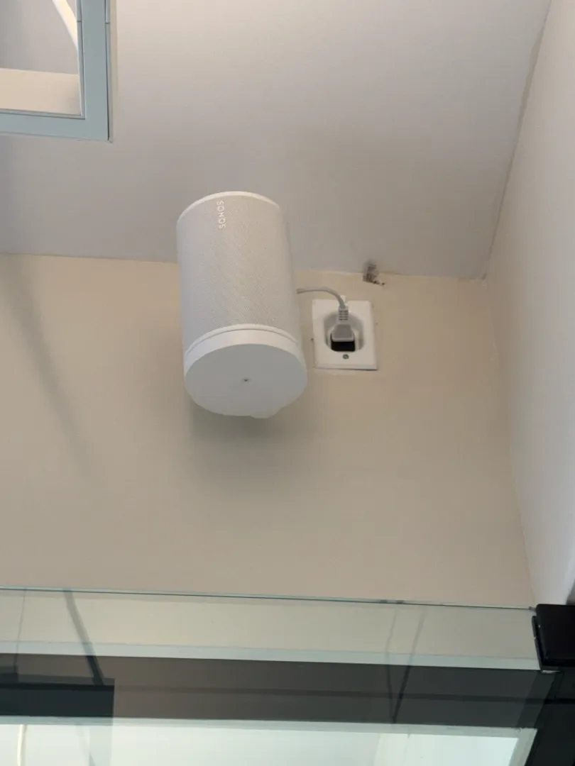 A white Sonos speaker mounted high on the wall next to its outlet, above a glass partition.