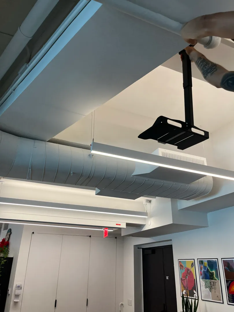 A technician fitting a black projector mount to a pipe under an open ceiling.