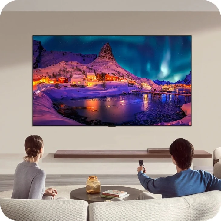 LG G6 OLED TV mounted on a living room wall above a floating shelf, showing a snowy aurora borealis scene, with a couple watching from the sofa