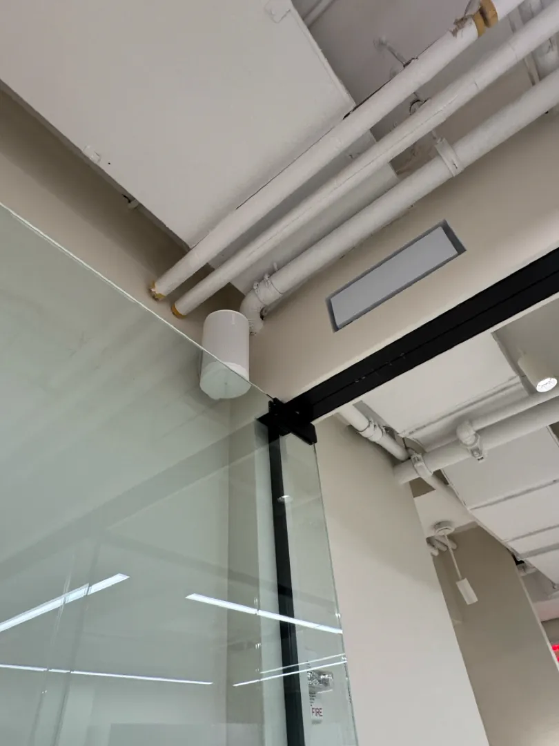 A white speaker mounted near the ceiling pipes above a glass office wall.