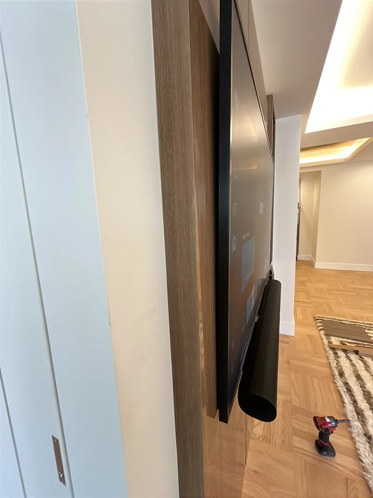 A TV is mounted on a wood paneled wall with its screen on, a power drill resting on the parquet floor nearby and a rug and ha