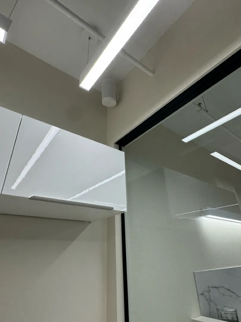 A small white speaker mounted high in the corner of an office ceiling beside a glass partition.