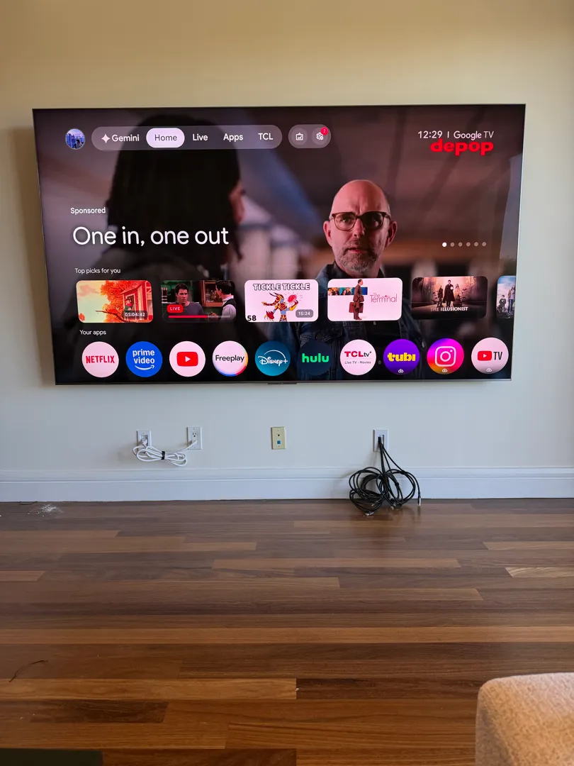 A very large wall-mounted TV showing its home screen above a baseboard and two outlets.