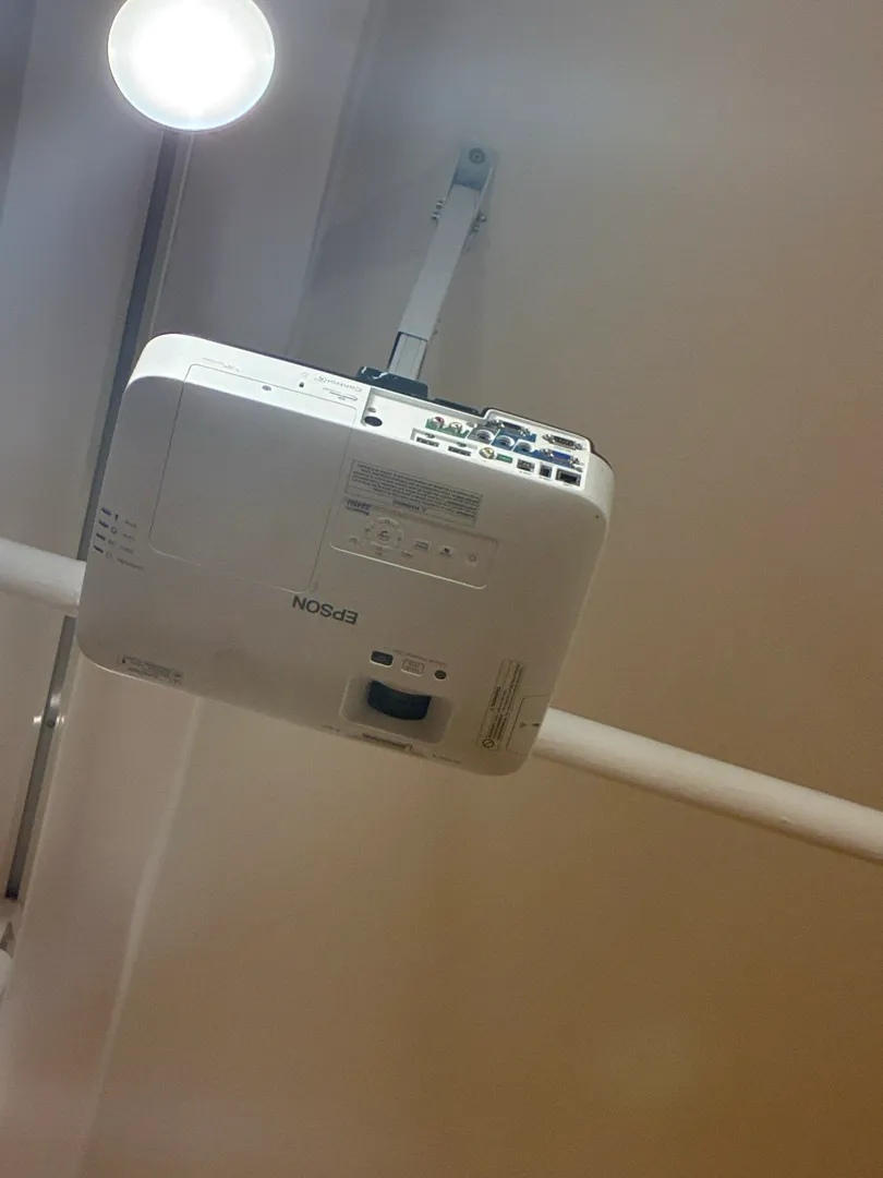 A white projector on a ceiling arm seen from below, with a pipe and a light.