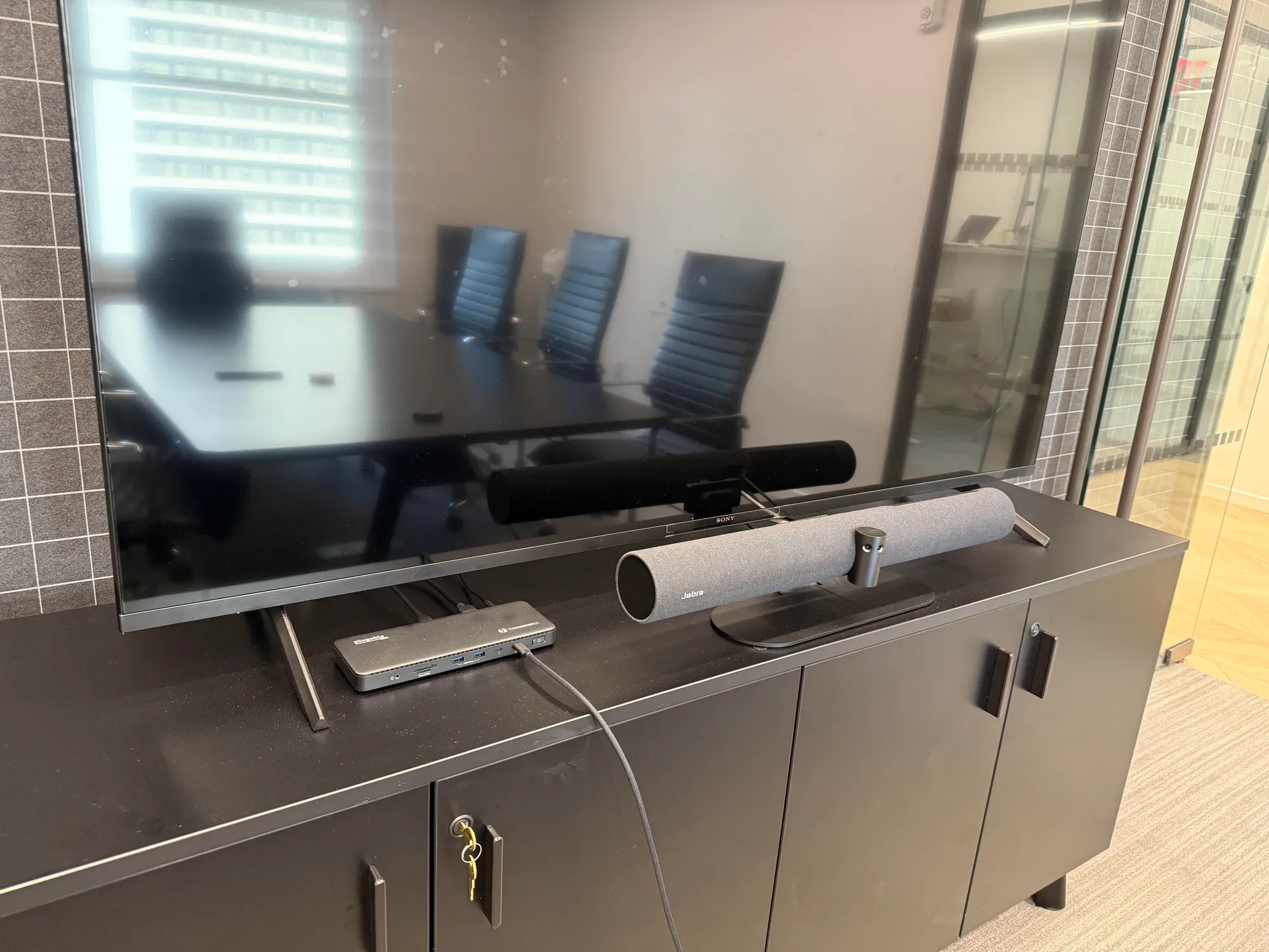 A TV still on its own tabletop stand sits on a cabinet in a conference room, with a Sony soundbar and a Jabra speaker bar bes