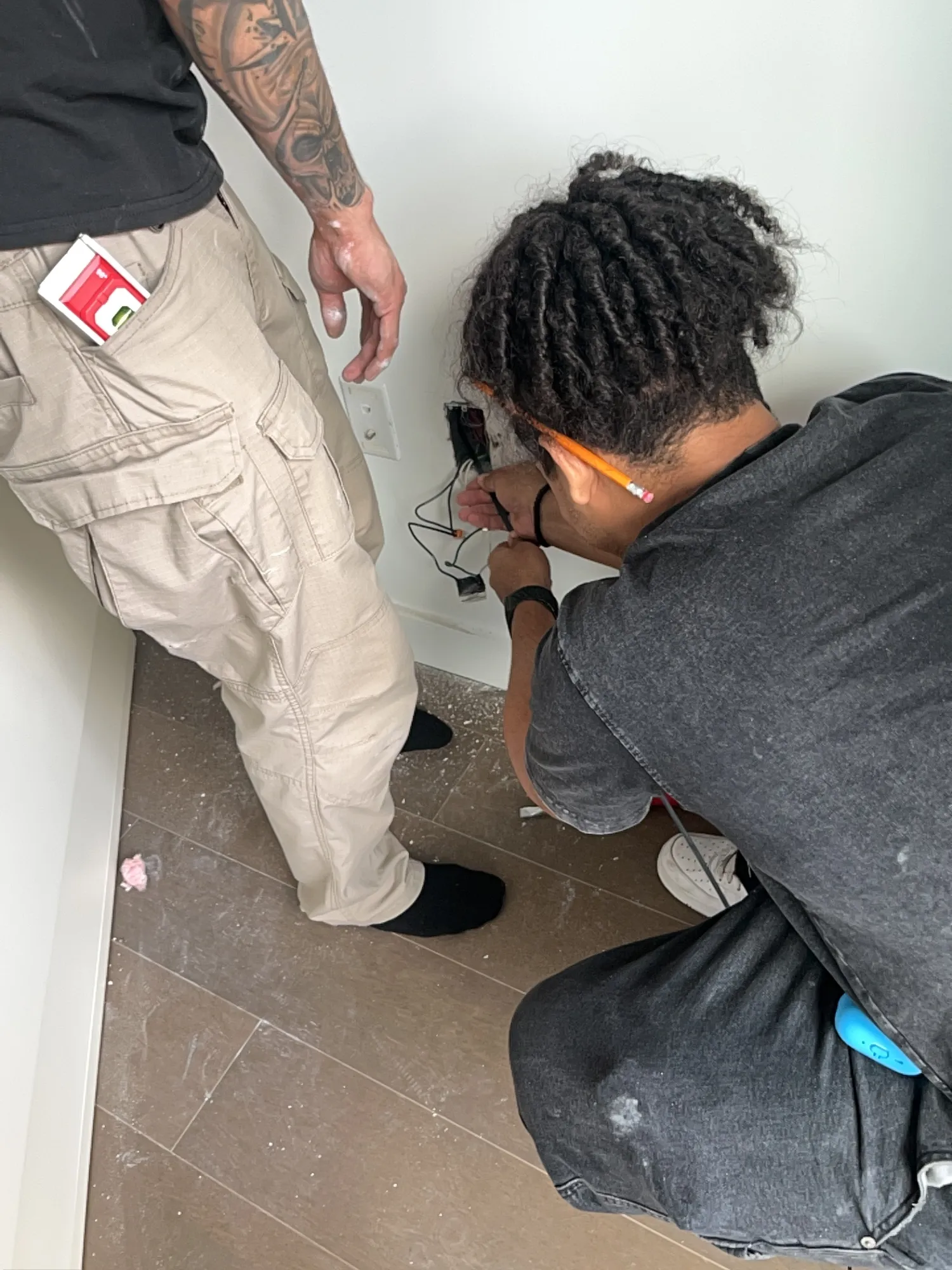 Two crew members working at an open electrical outlet box in the wall, wires visible.