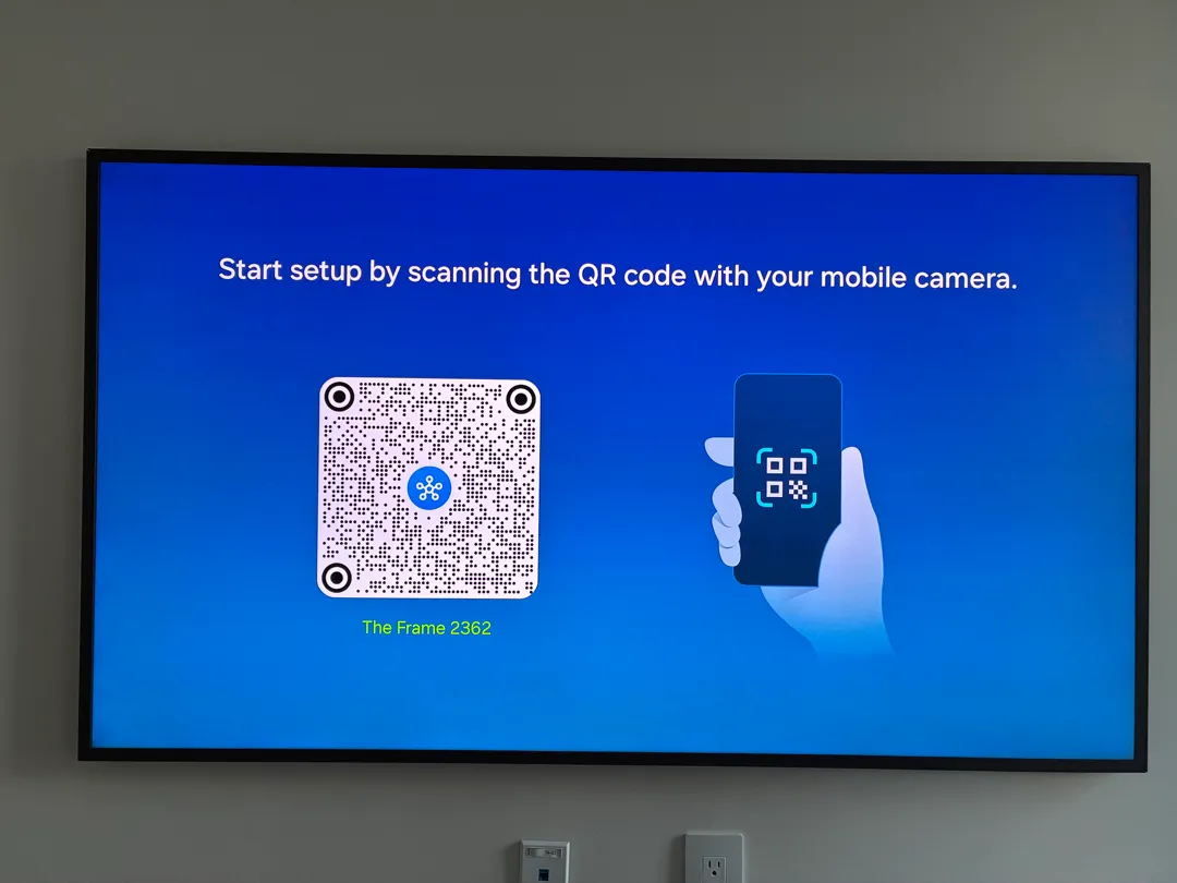 A Samsung Frame TV hanging flat on a wall above a cable plate and an outlet showing a setup screen.