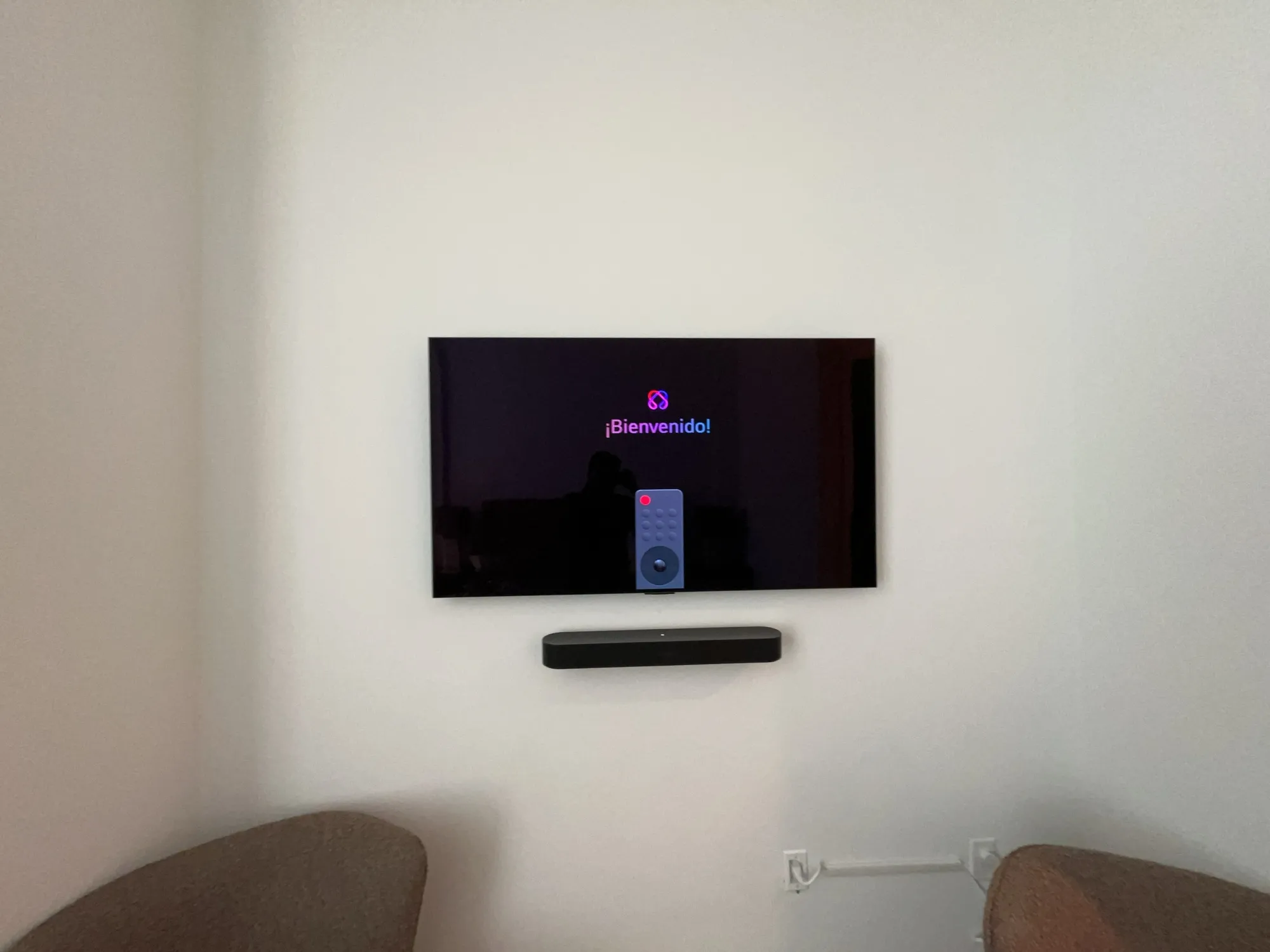 A wall-mounted TV showing its welcome screen, with a soundbar beneath and chairs in front.