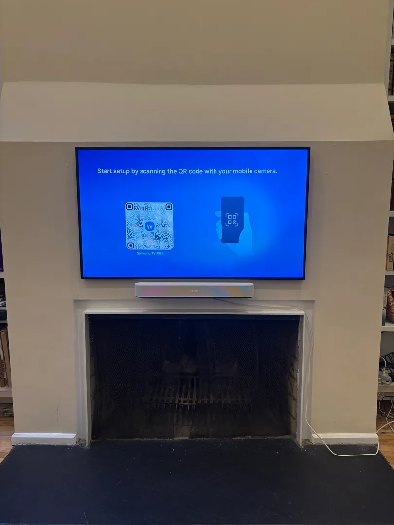 A TV showing a setup screen hangs flat on the chimney wall above a fireplace with a white soundbar beneath it.