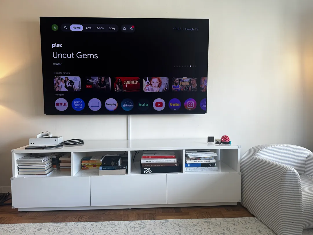 A wall-mounted TV shows a streaming home screen above a white media console with books and a striped armchair nearby.