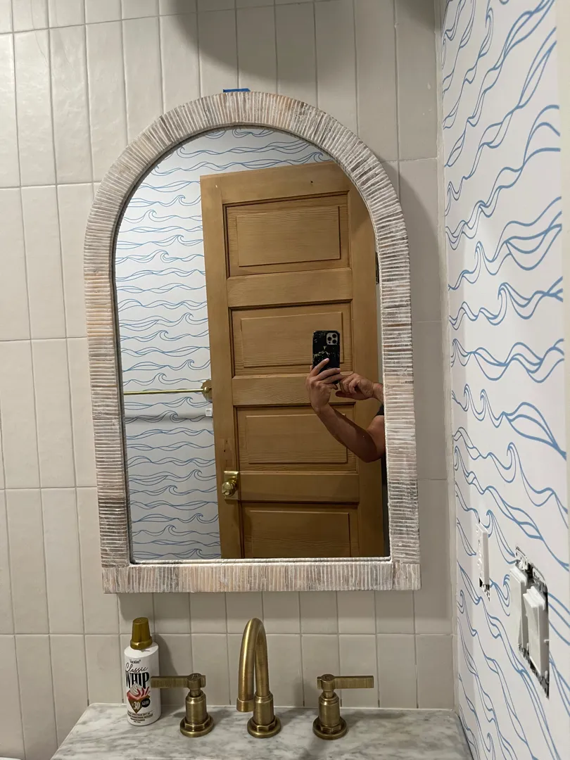 A large arched mirror mounted above a bathroom vanity with brass fixtures and patterned wallpaper.