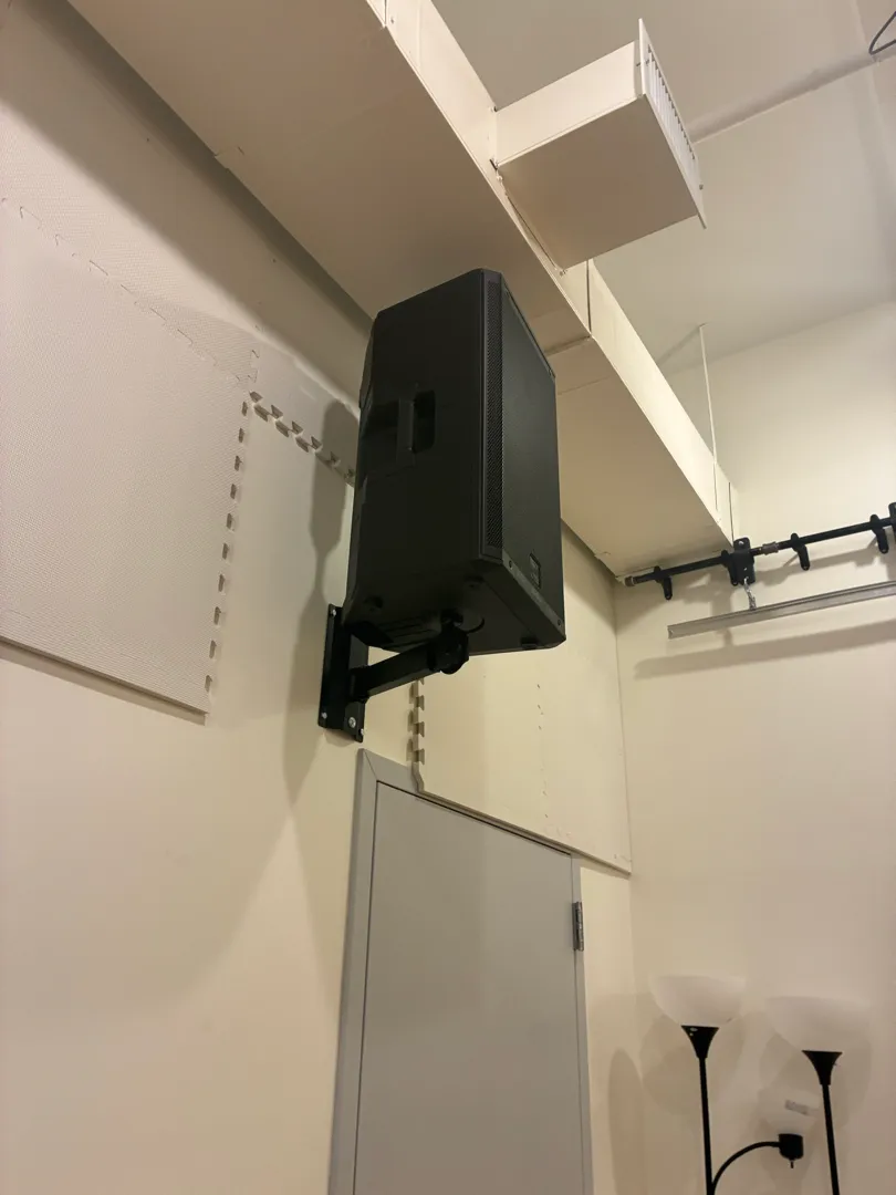 A black speaker on a wall bracket above a gray door, with a curtain rail and two floor lamps nearby.