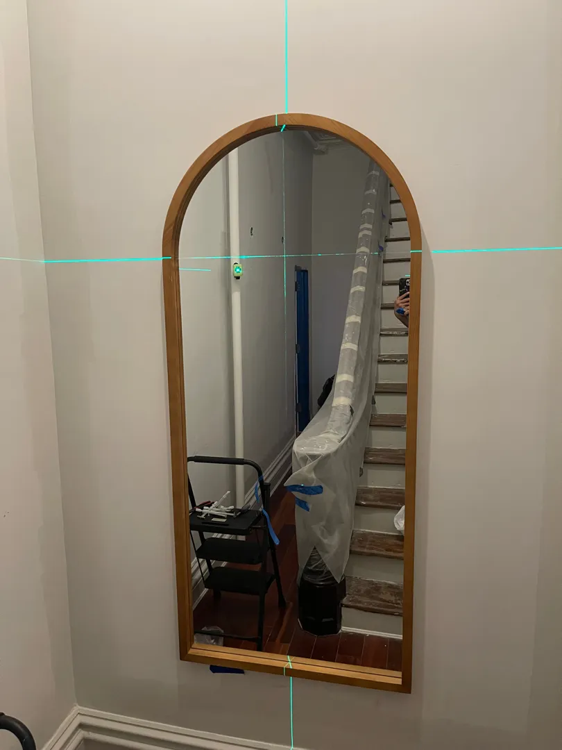 A large arched wood-framed mirror being leveled near a staircase, laser guide lines visible.