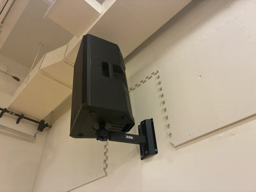 A black speaker hanging from a black wall bracket high on a cream wall with foam tiles.