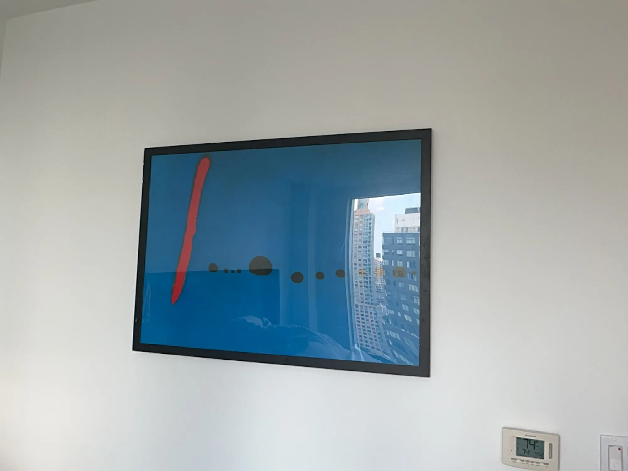 A framed abstract print, hung level on the wall, reflecting a city skyline through a nearby window.