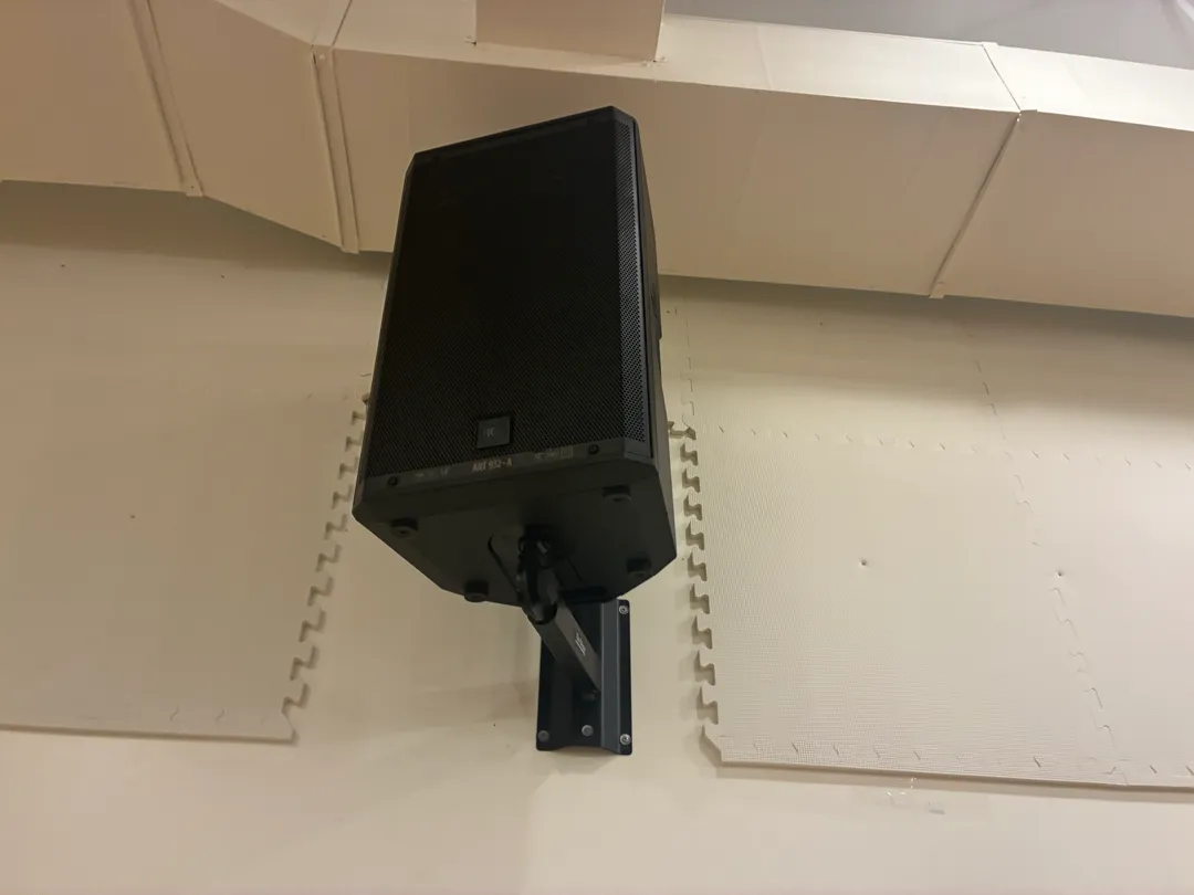 A black speaker on a wall bracket under a ceiling duct, with foam tiles around it.