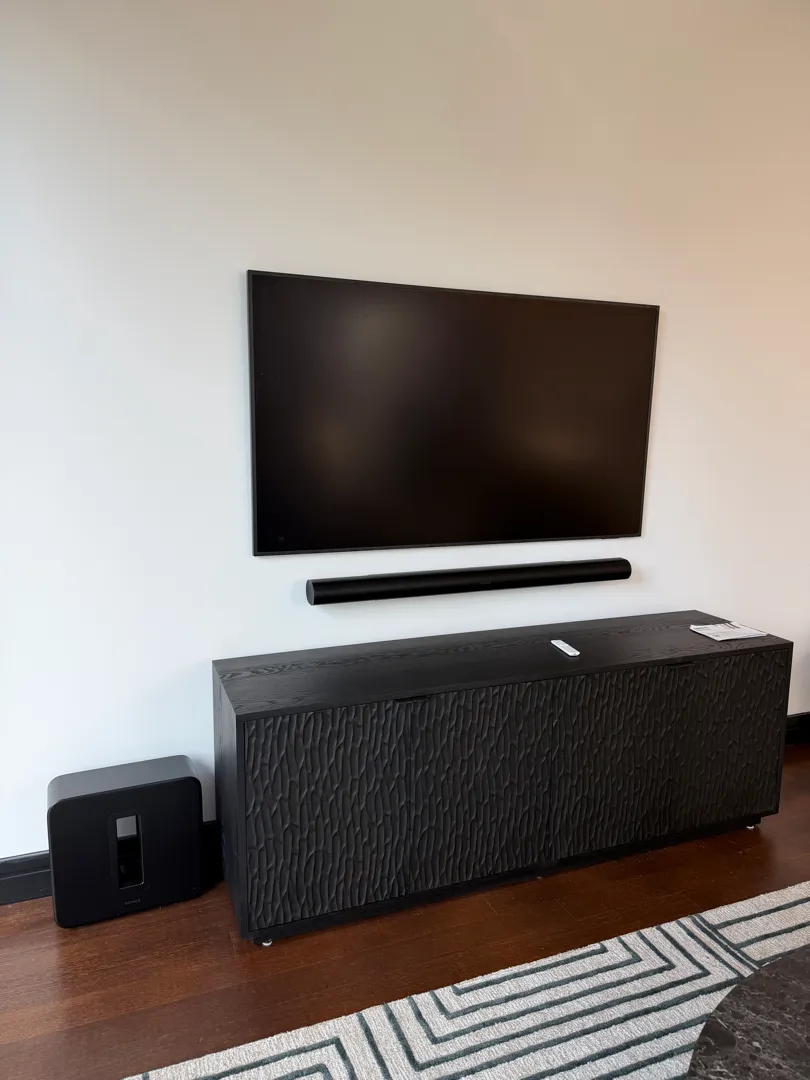 TV and soundbar are mounted on a wall above a black console, with a Sonos subwoofer on the floor beside it.