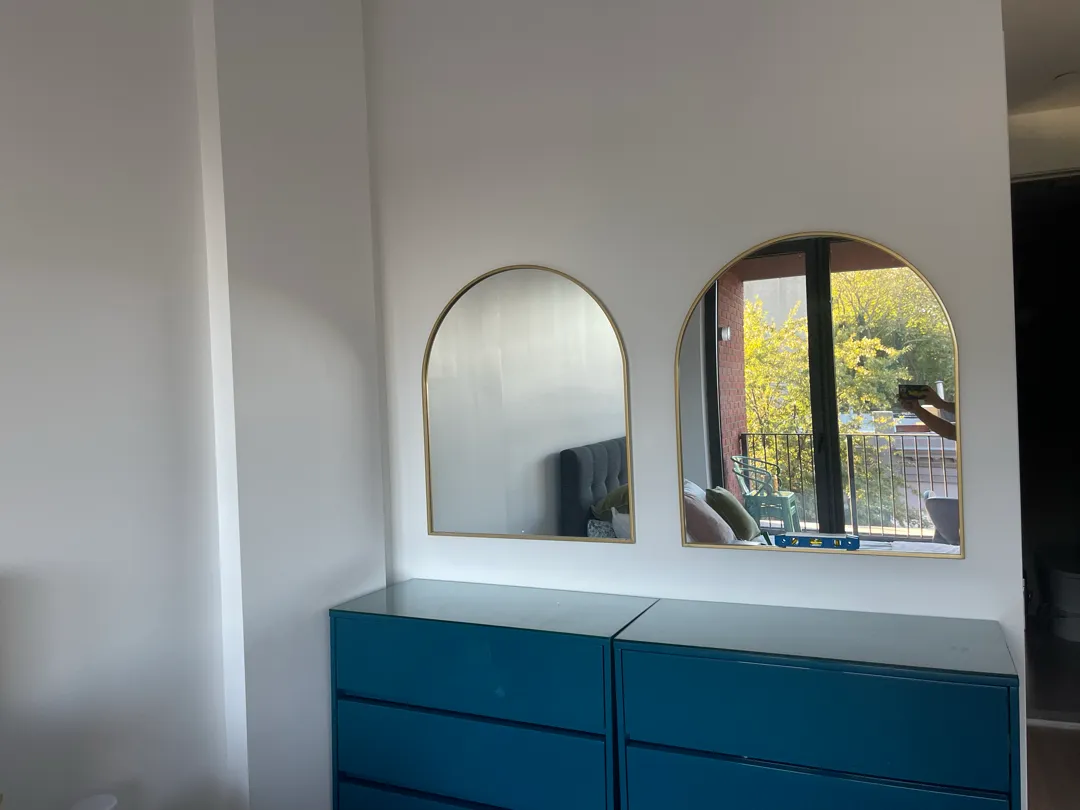 Two arched mirrors with brass frames hanging side by side above two teal dressers, with a level on the dresser top.
