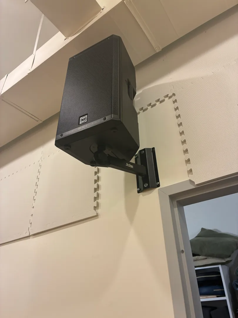 A black powered speaker on a wall bracket beside a doorway, with foam tiles on the wall.