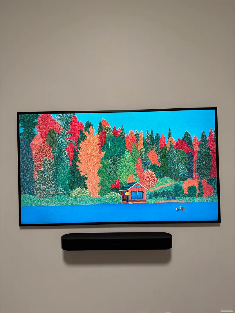 A Samsung Frame Pro TV showing a colourful forest painting, mounted flush on a grey wall above a compact black soundbar.