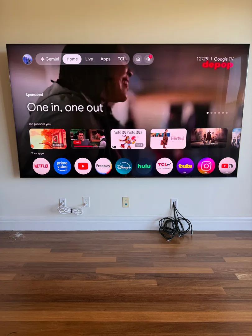 A very large TV hanging flat on a cream wall above two outlets and a wood floor.