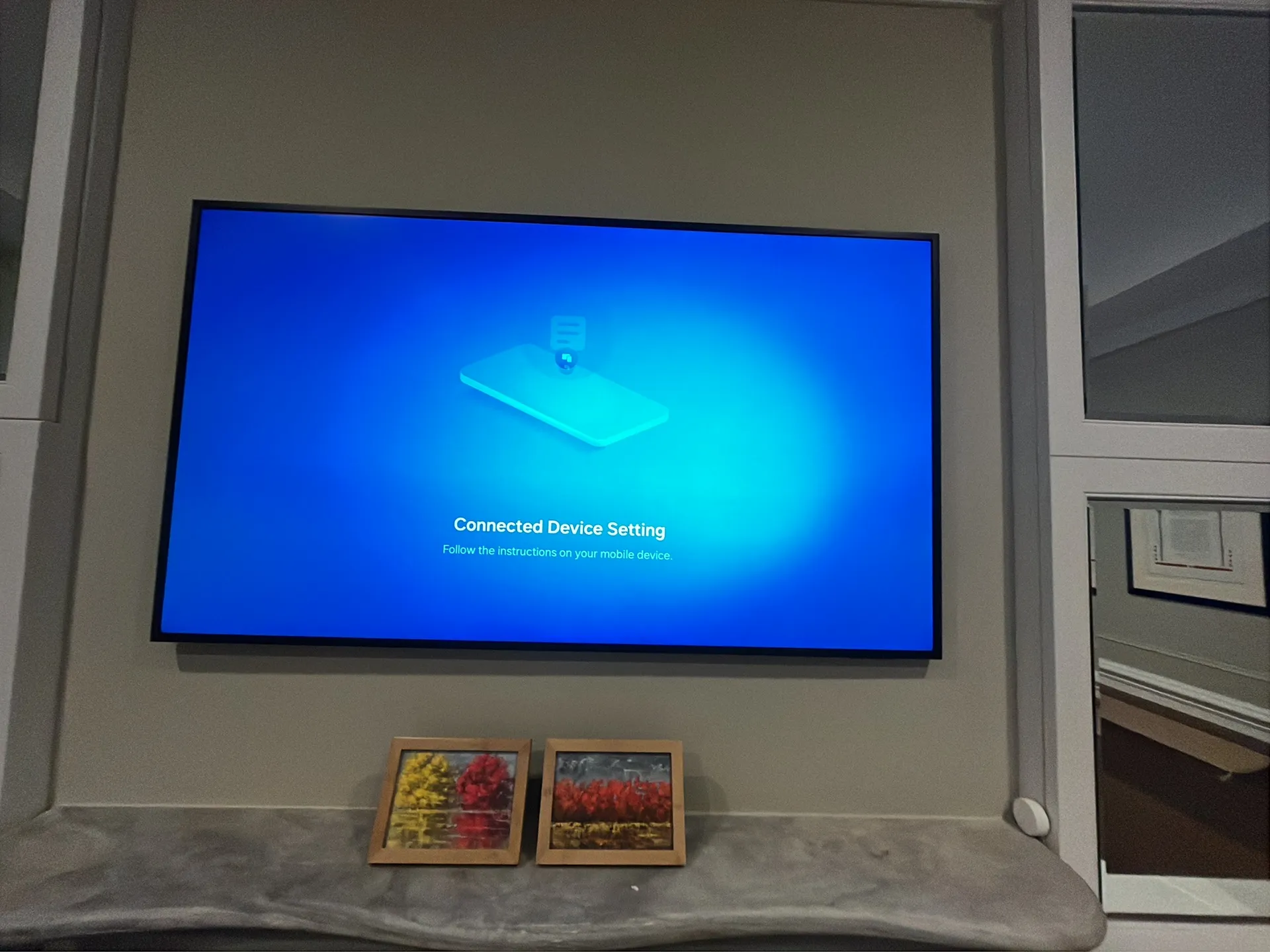 A mounted TV displays a connected device setup screen, above a stone ledge holding two small framed paintings.