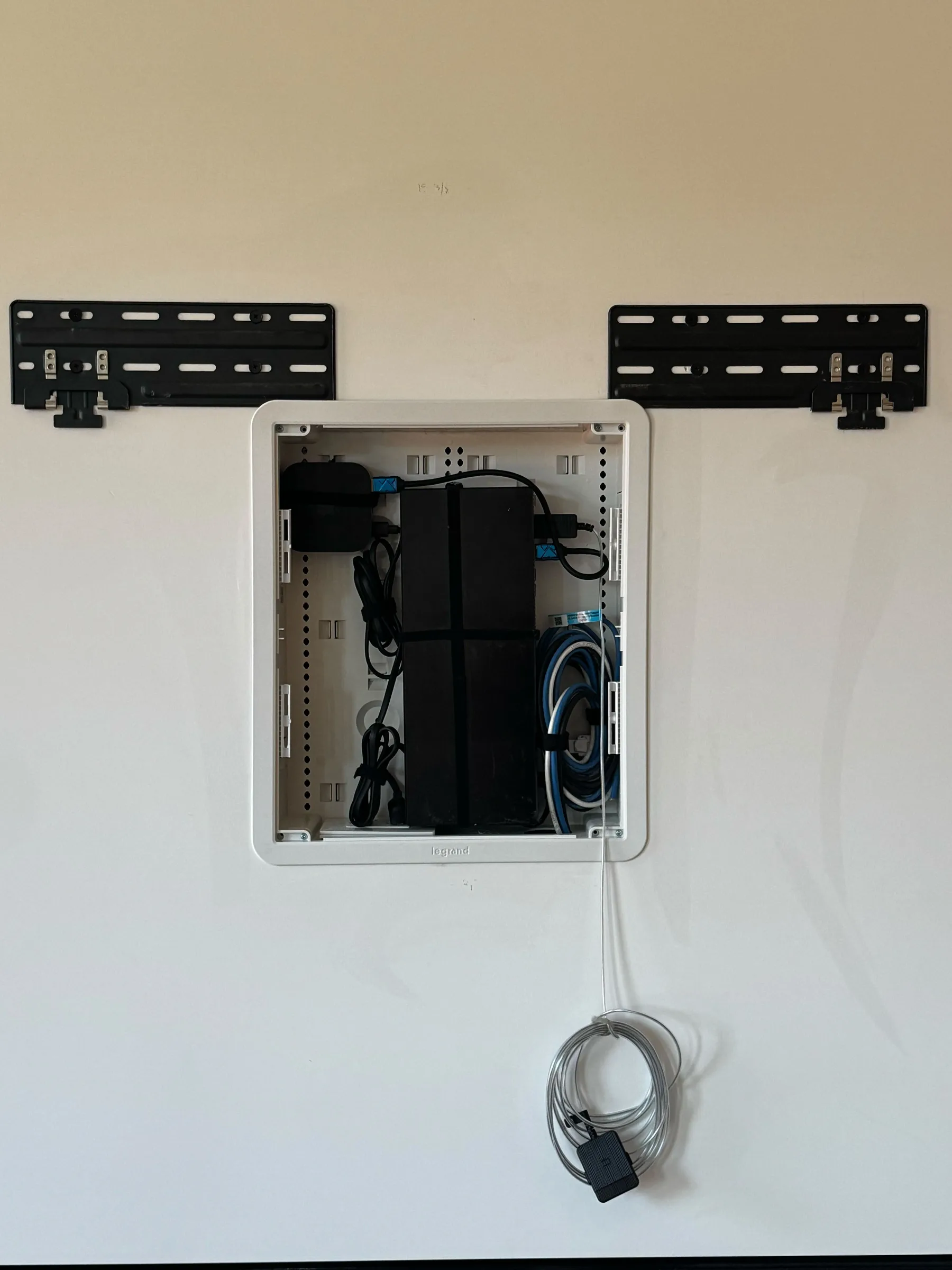 An open in-wall AV cable box with equipment inside, flanked by two black TV mount brackets on a wall.