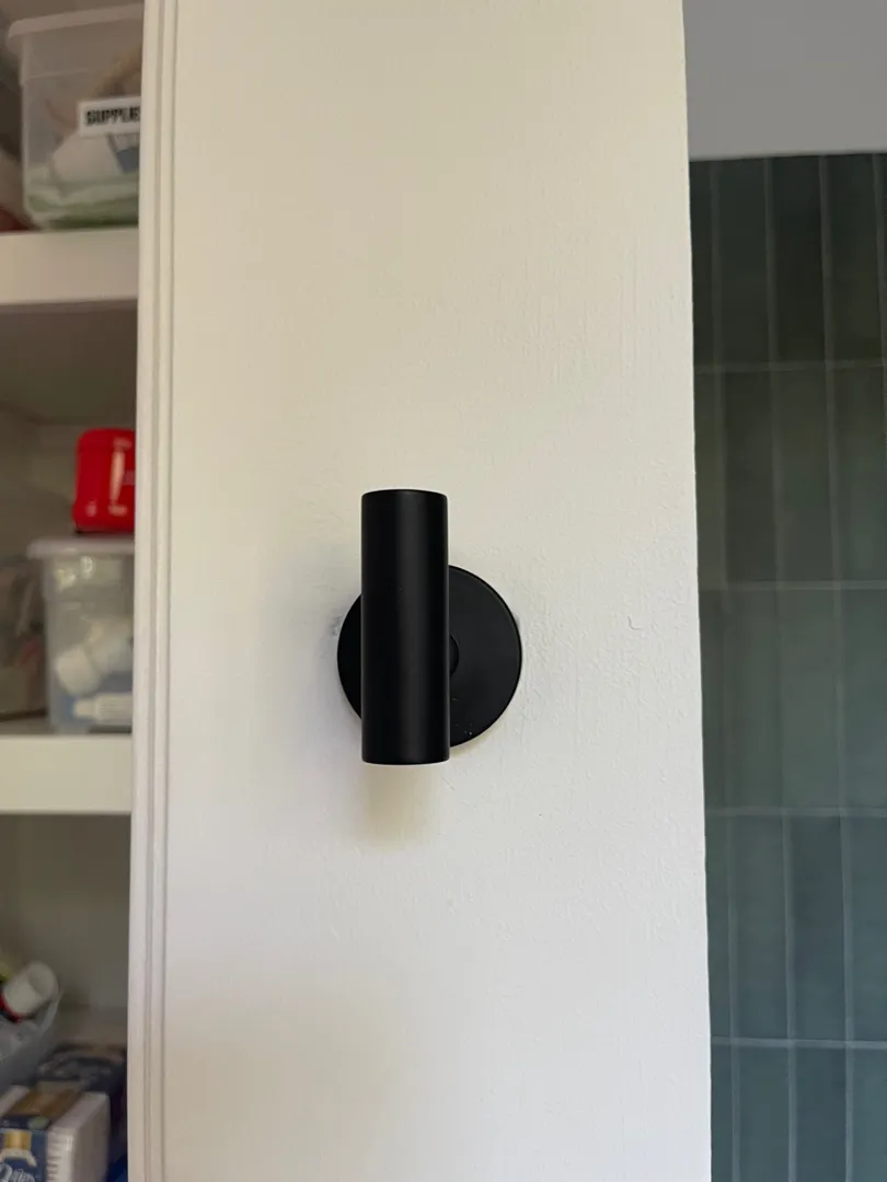 A black wall hook mounted on a white wall near a bathroom.