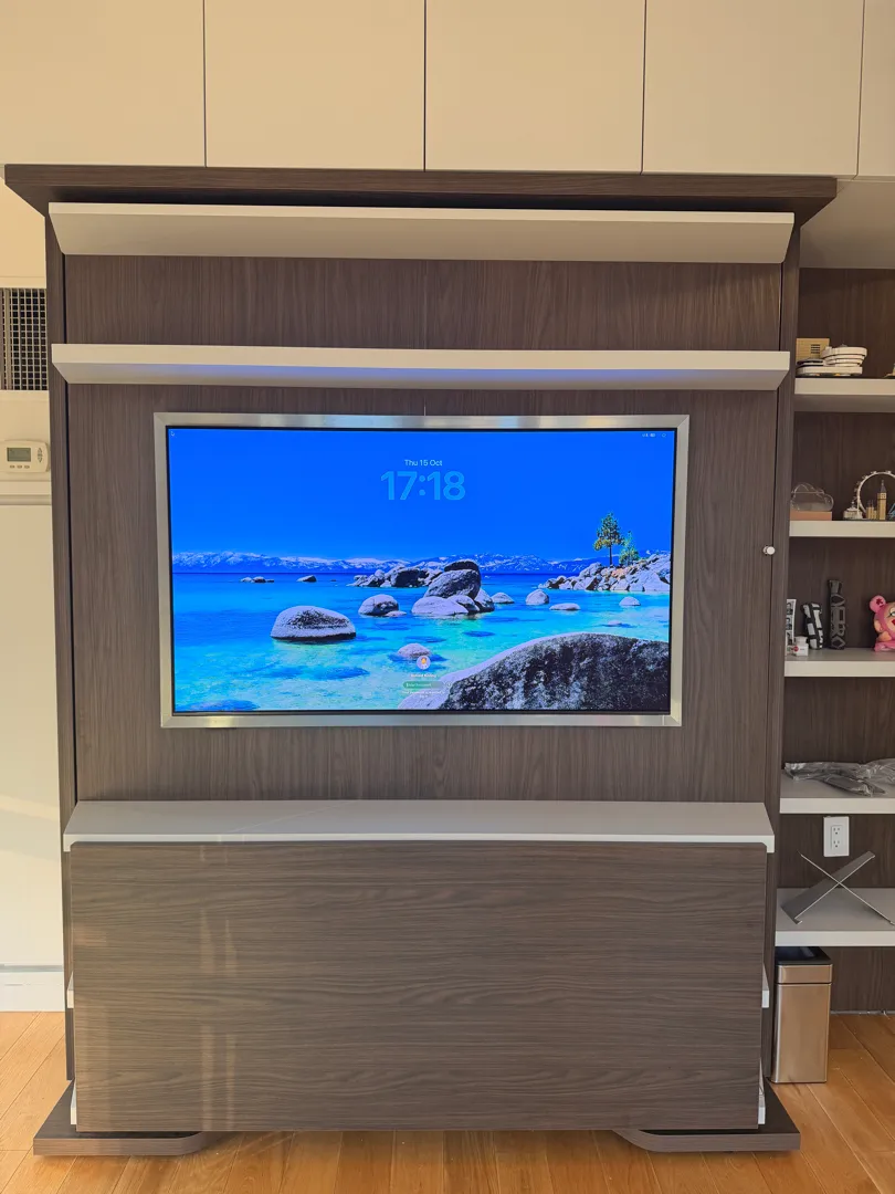 A TV showing a lock screen is mounted inside a wood cabinet unit with a lift mechanism, flanked by open shelving with decor.