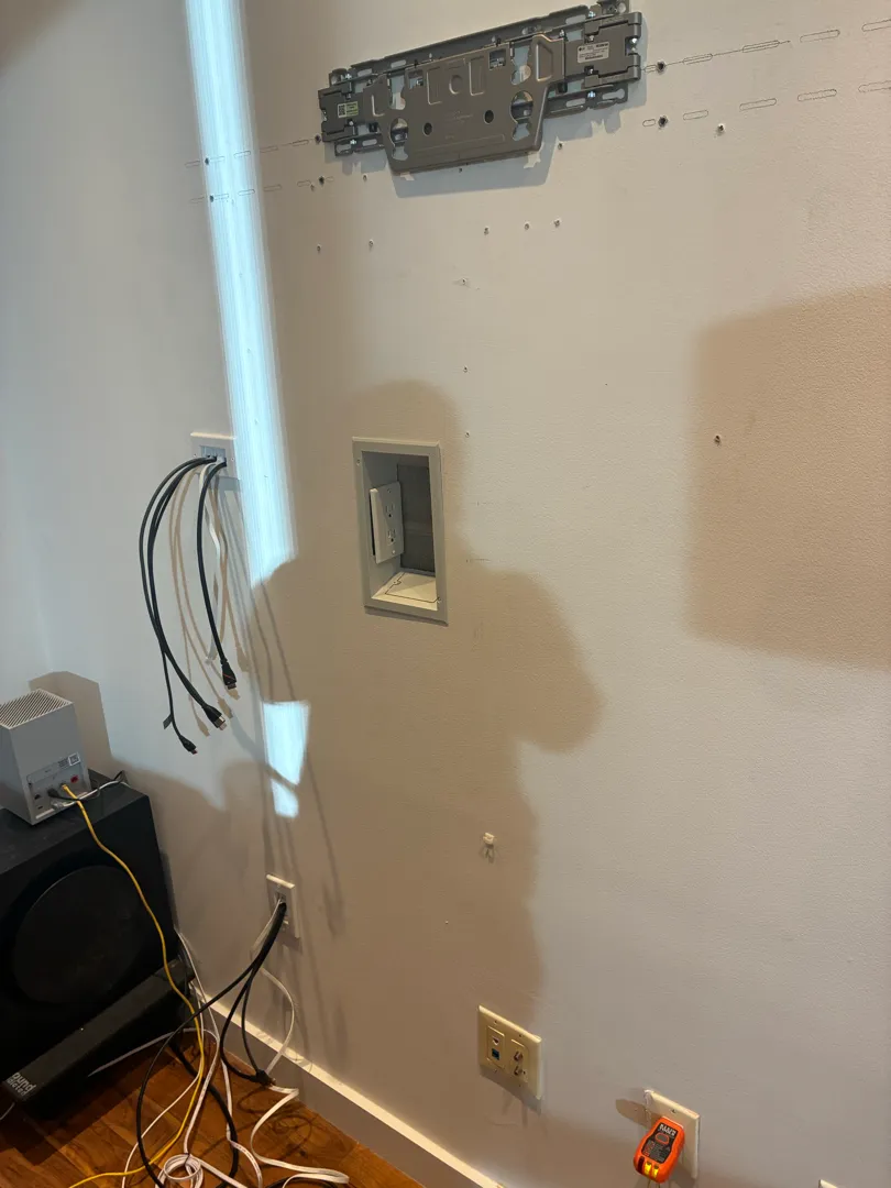 A metal TV mounting plate on the wall above a recessed outlet box with cables nearby.