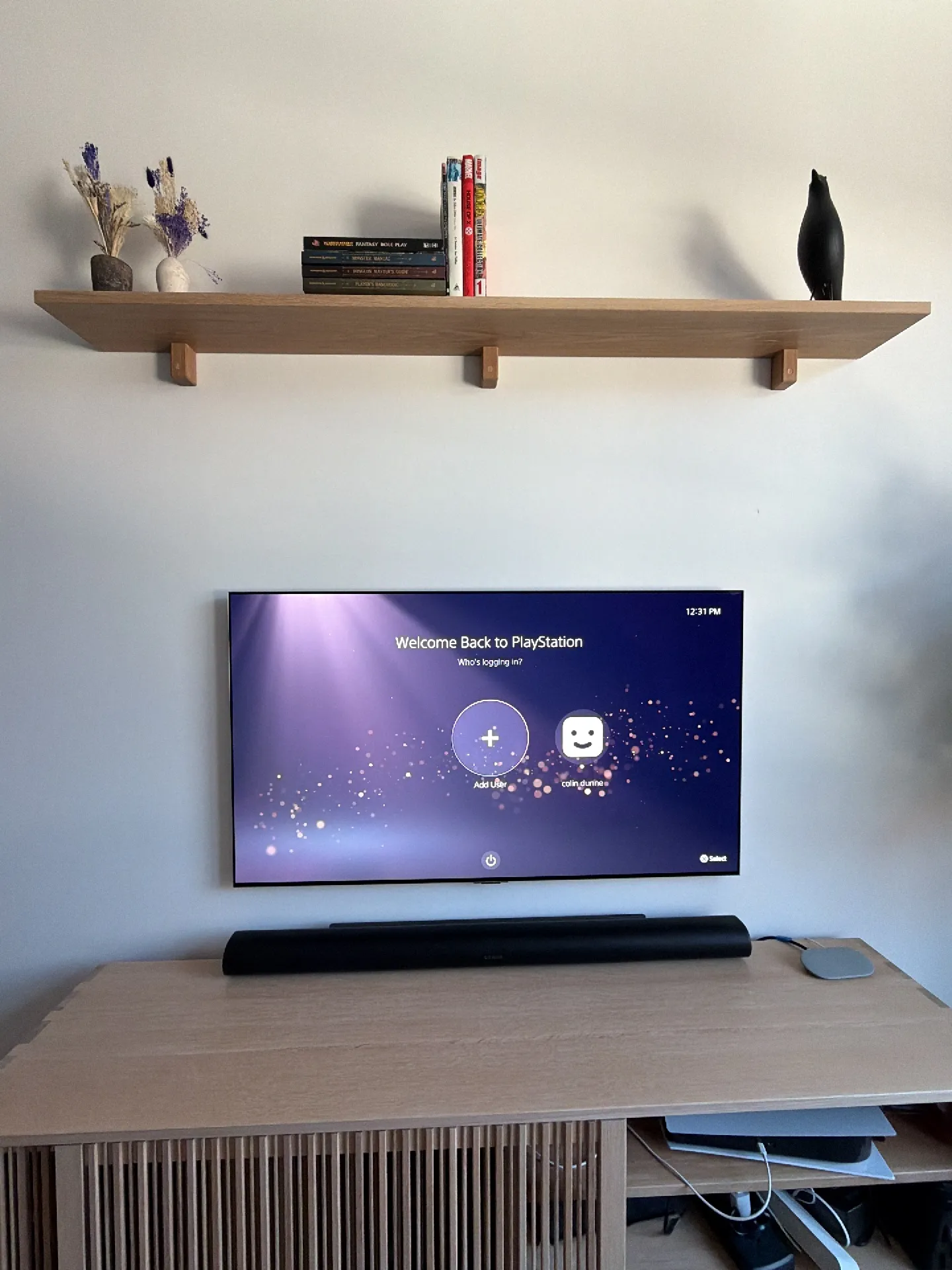 A wall-mounted TV shows a PlayStation login screen below a wood shelf holding books and decor, with a soundbar on a console u