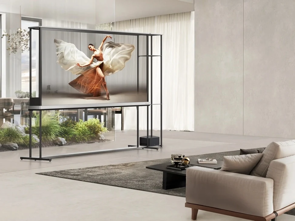 LG SIGNATURE OLED T in a bright living room with sheer curtains and a sofa, displaying a ballet dancer mid-spin, the transparent frame letting the room show through around the image