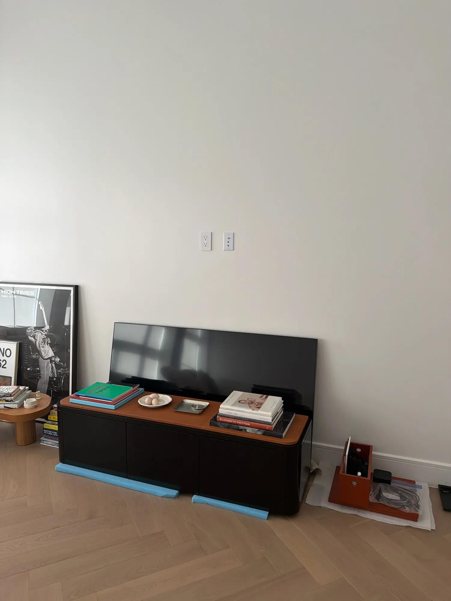 An unmounted TV leaning on a media console below two wall outlets, mounting hardware in packaging.