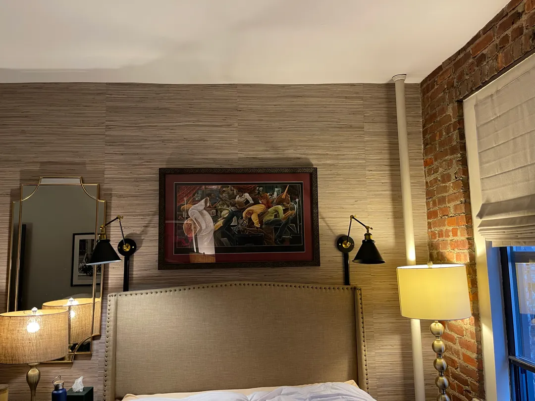 A large framed print on a grasscloth wall above a headboard, between two black swing-arm wall lights.