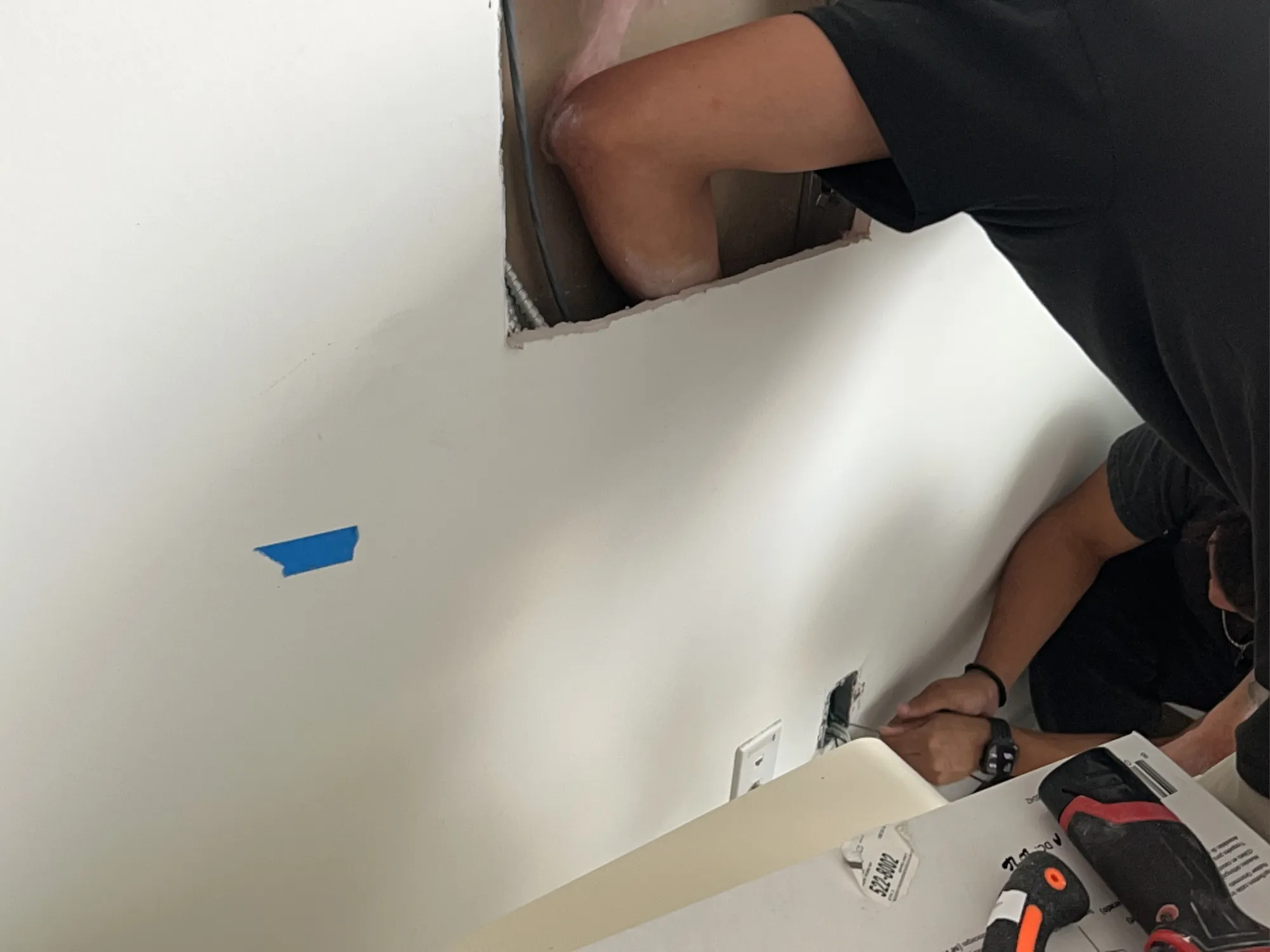 A crew member reaching into a cut-open wall cavity while another works at an open outlet box with a screwdriver.