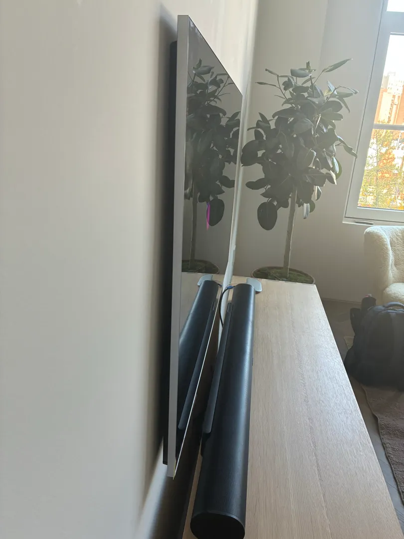 A TV is mounted on the wall at an angle showing a reflection of a potted plant, with a soundbar on a wood console below.
