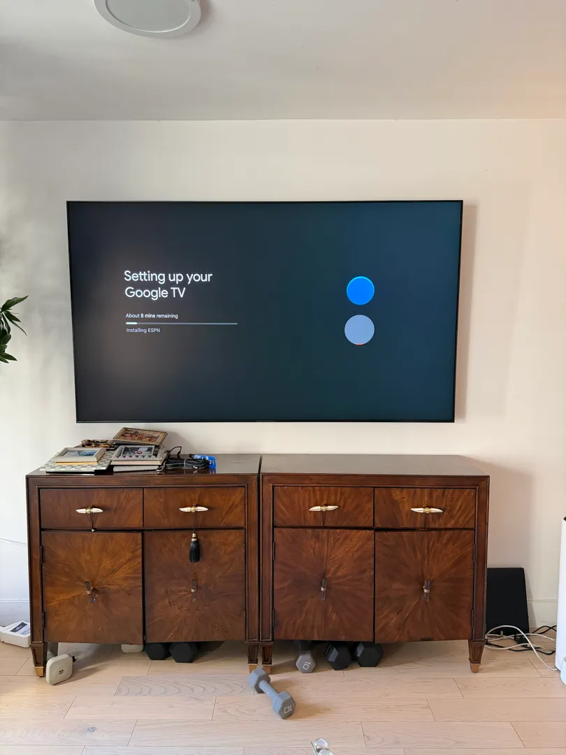 A wide view of the mounted TV showing a Google TV setup screen, with a speaker and dumbbells on the floor nearby.