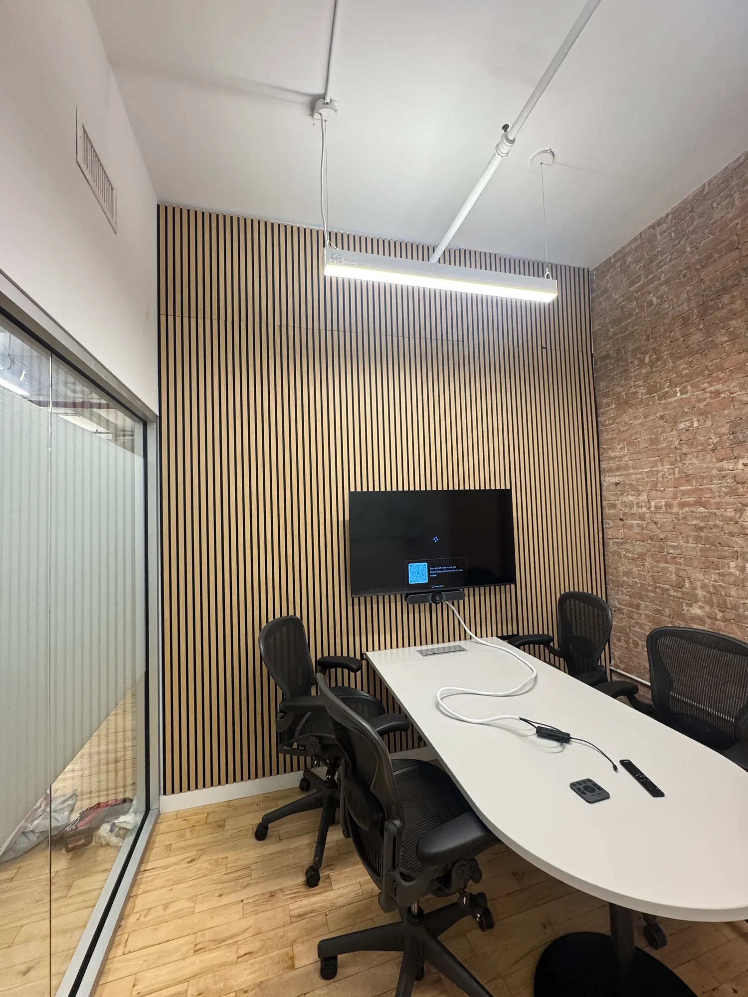 A room with an exposed brick wall and a TV mounted on a wood panel wall, an oval table and chairs in front.